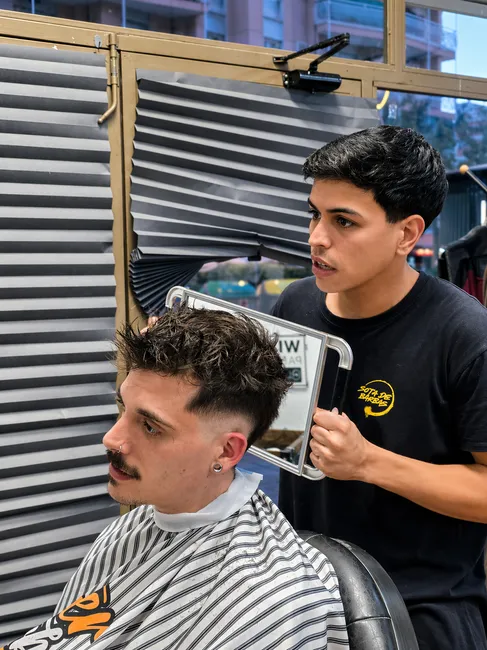 Un barbero muestra en un espejo de mano el corte de un cliente sentado en el sillón.